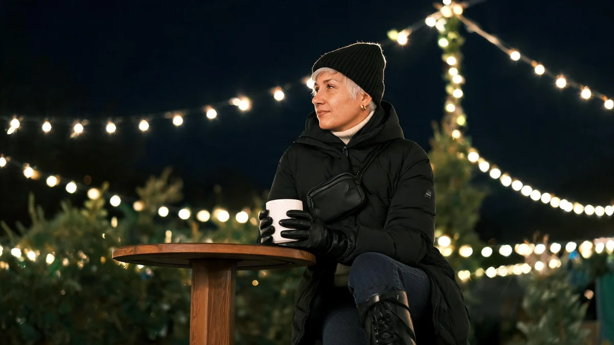Frau im knöchellangen schwarzen Daunenmantel mit cremefarbenem Rollkragen, Jeans und Schnürboots an einem Stehtisch auf dem Weihnachtsmarkt