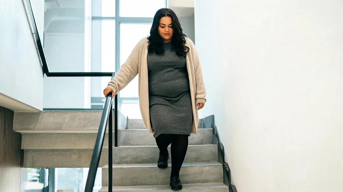 Plus-size woman in her thirties wearing a charcoal knee-length knit dress, an open oatmeal longline cardigan, black tights and loafers on an office staircase