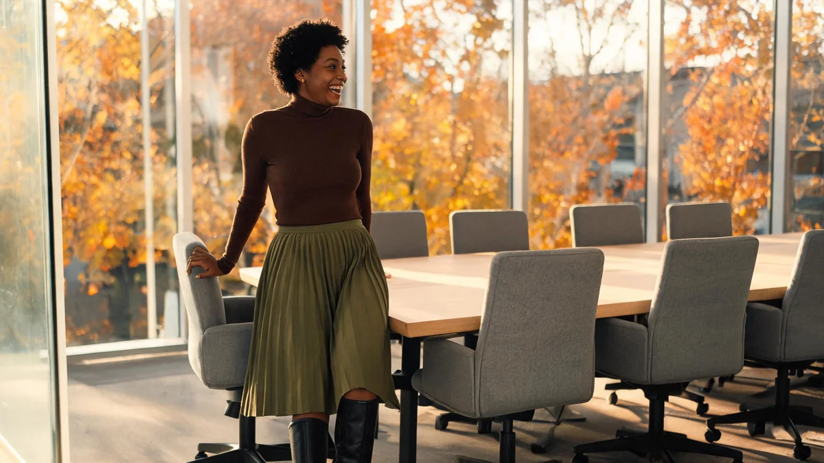 Young woman with short natural hair in a brown ribbed turtleneck, pleated olive midi skirt and black knee-high boots in a glass-walled meeting room