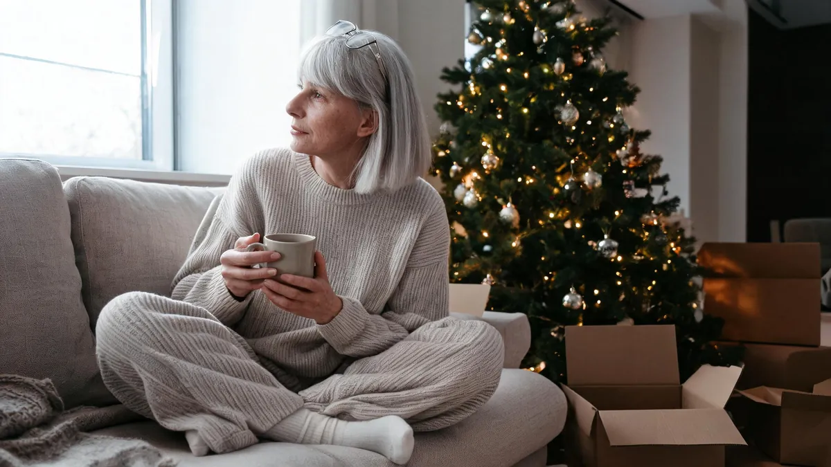Woman in her fifties in a matching oatmeal knit set and wool socks, sitting on a sofa beside a decorated Christmas tree with a mug of coffee