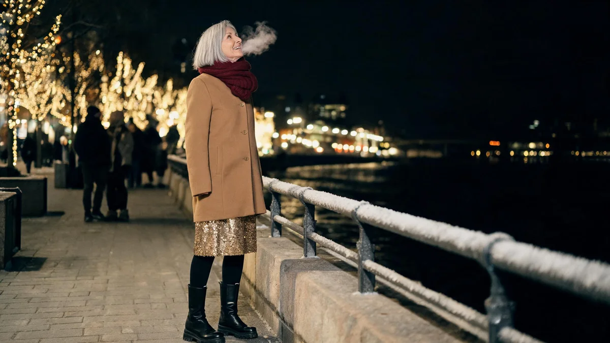 Woman in her fifties in a long camel coat over a gold sequin midi dress, black knee boots and a burgundy scarf on a cold night