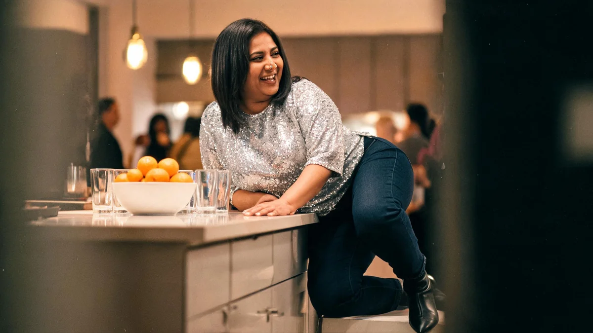 Woman in a silver sequin top, dark straight-leg jeans and black ankle boots leaning on a kitchen counter at a house party
