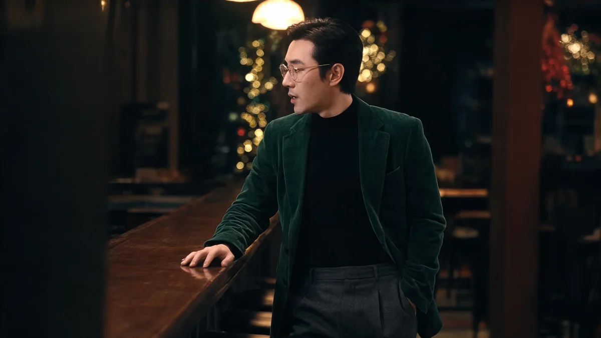 Man in a bottle-green velvet blazer, black roll-neck and charcoal trousers at a dimly lit restaurant bar