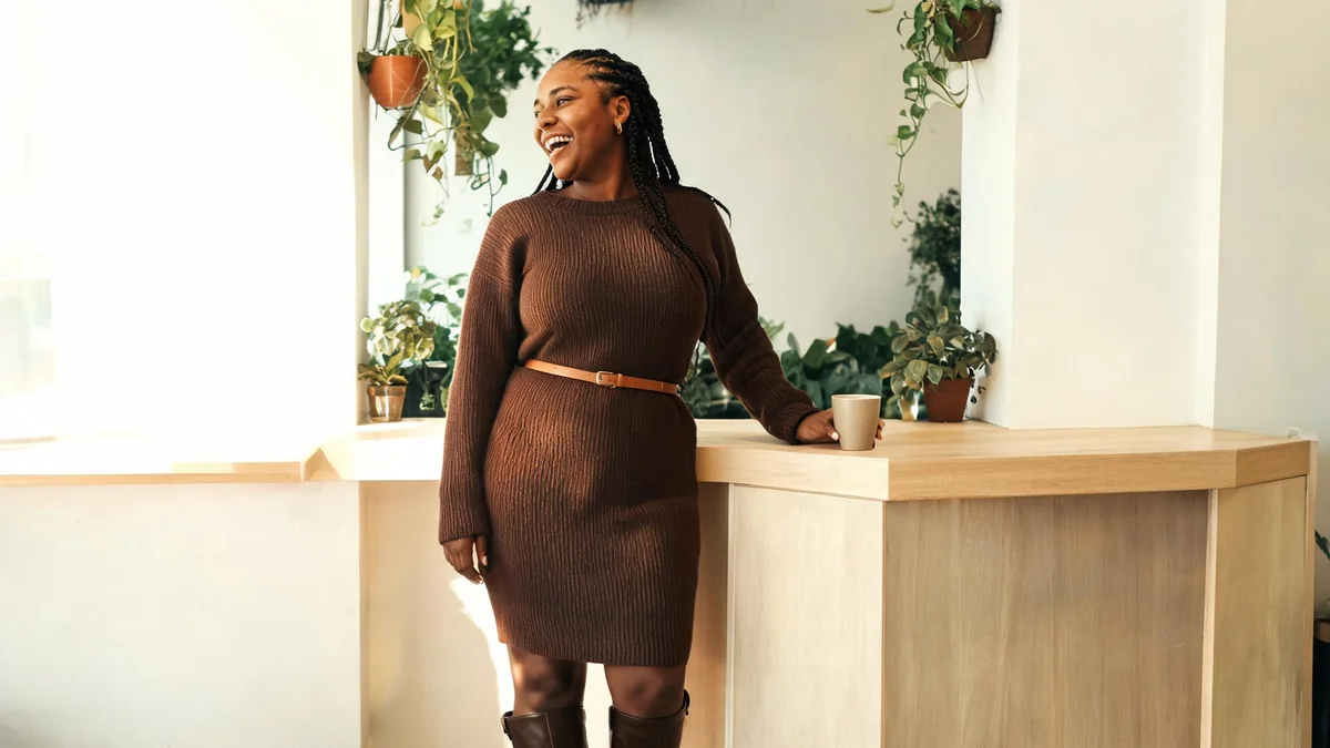 Woman with box braids in a chocolate-brown ribbed sweater dress, thin cognac belt and brown knee-high boots in a café