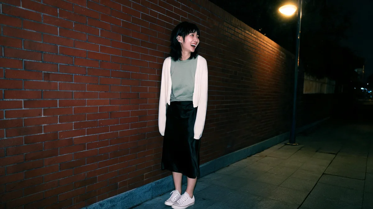 Young woman in a faded sage tee, black satin slip skirt, open cream cardigan and white canvas sneakers outside a small music club at night