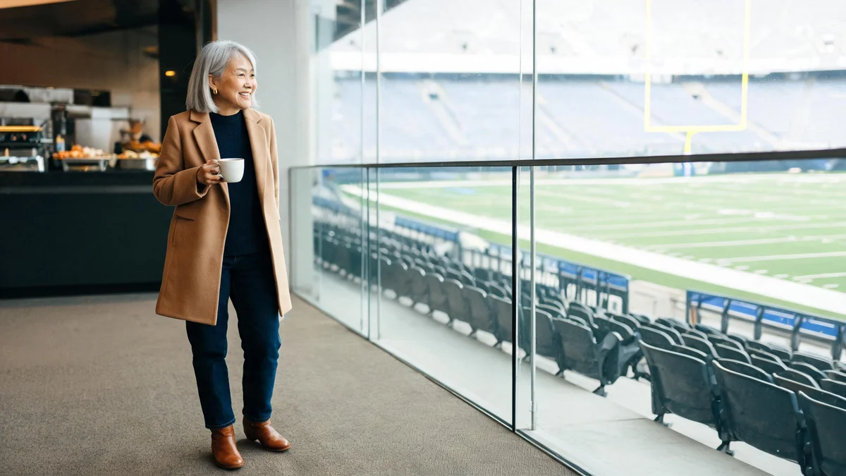 Woman in a stadium suite with a camel wool coat over her shoulders, navy merino sweater, dark jeans and cognac ankle boots, standing at the glass above the field
