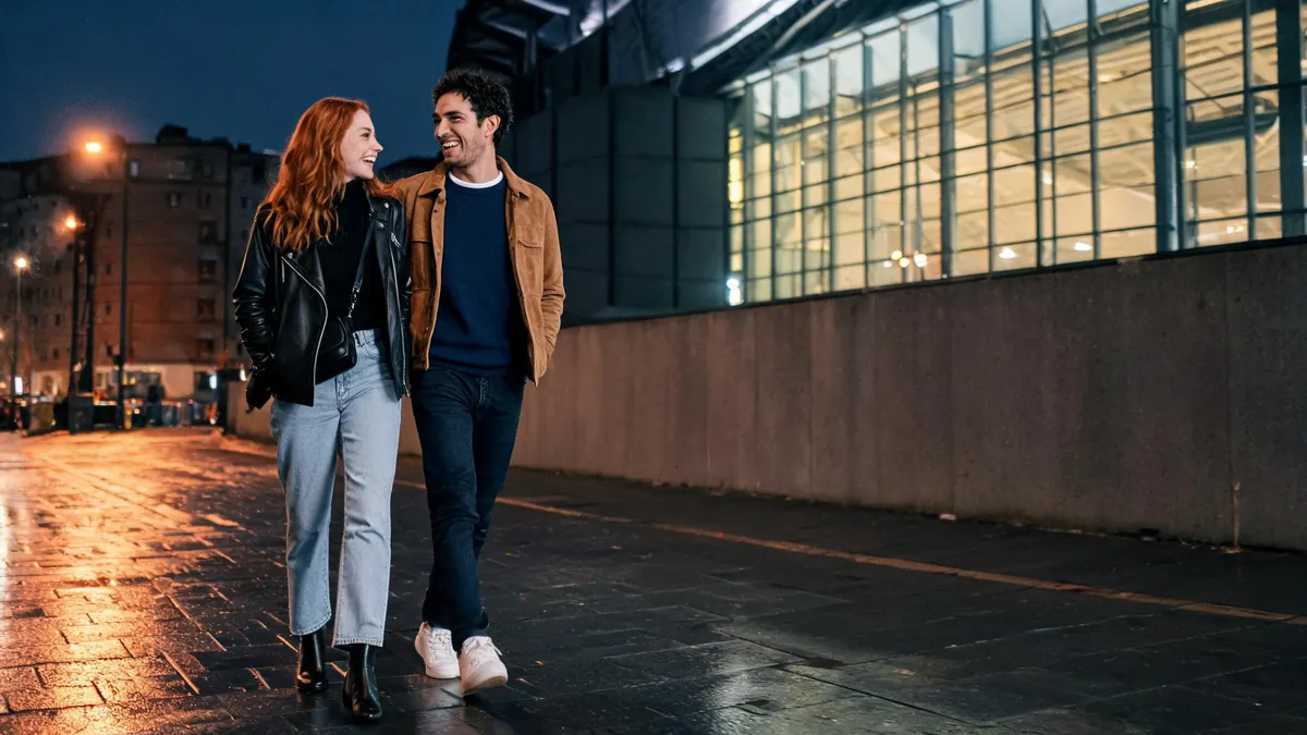 Couple walking outside an arena at night, she in a black turtleneck, leather jacket and light jeans, he in a navy crewneck and tan suede jacket
