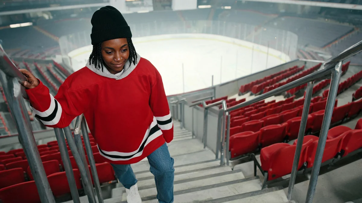 Woman in a plain red hockey jersey worn untucked over a grey hoodie, straight jeans and chunky sneakers on the stairs of an arena's upper level