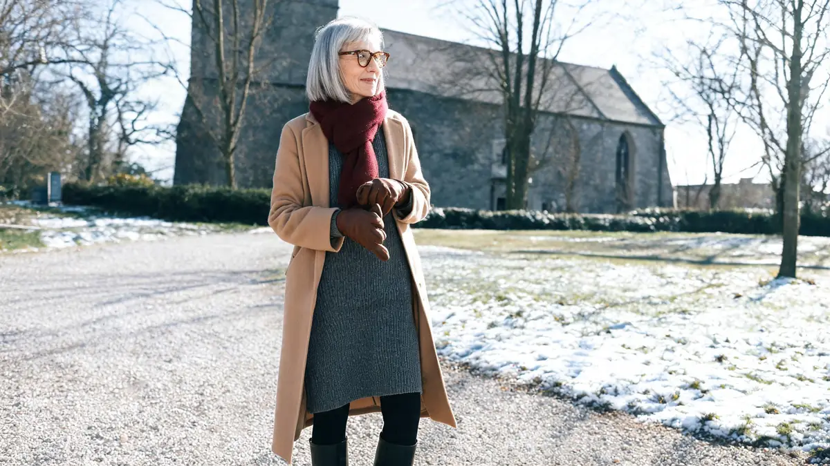 Woman in a charcoal sweater dress, black tights, knee boots and camel wool coat outside a stone church in winter