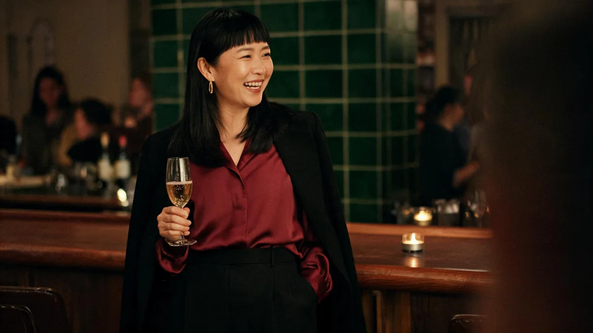 Woman in black tailored trousers, a burgundy satin blouse and an open black blazer with gold drop earrings at a wooden bar counter during after-work Christmas drinks