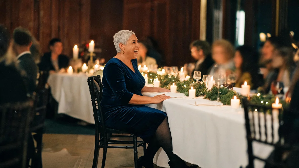 Woman in her fifties in a navy velvet wrap midi dress with three-quarter sleeves, sheer black tights and gold drop earrings at a candlelit restaurant table during a company Christmas dinner