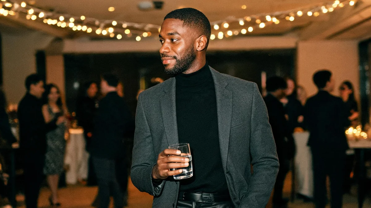 Man in his thirties in a charcoal wool suit with a black merino roll-neck and black Chelsea boots talking at a company Christmas party in a hotel function room