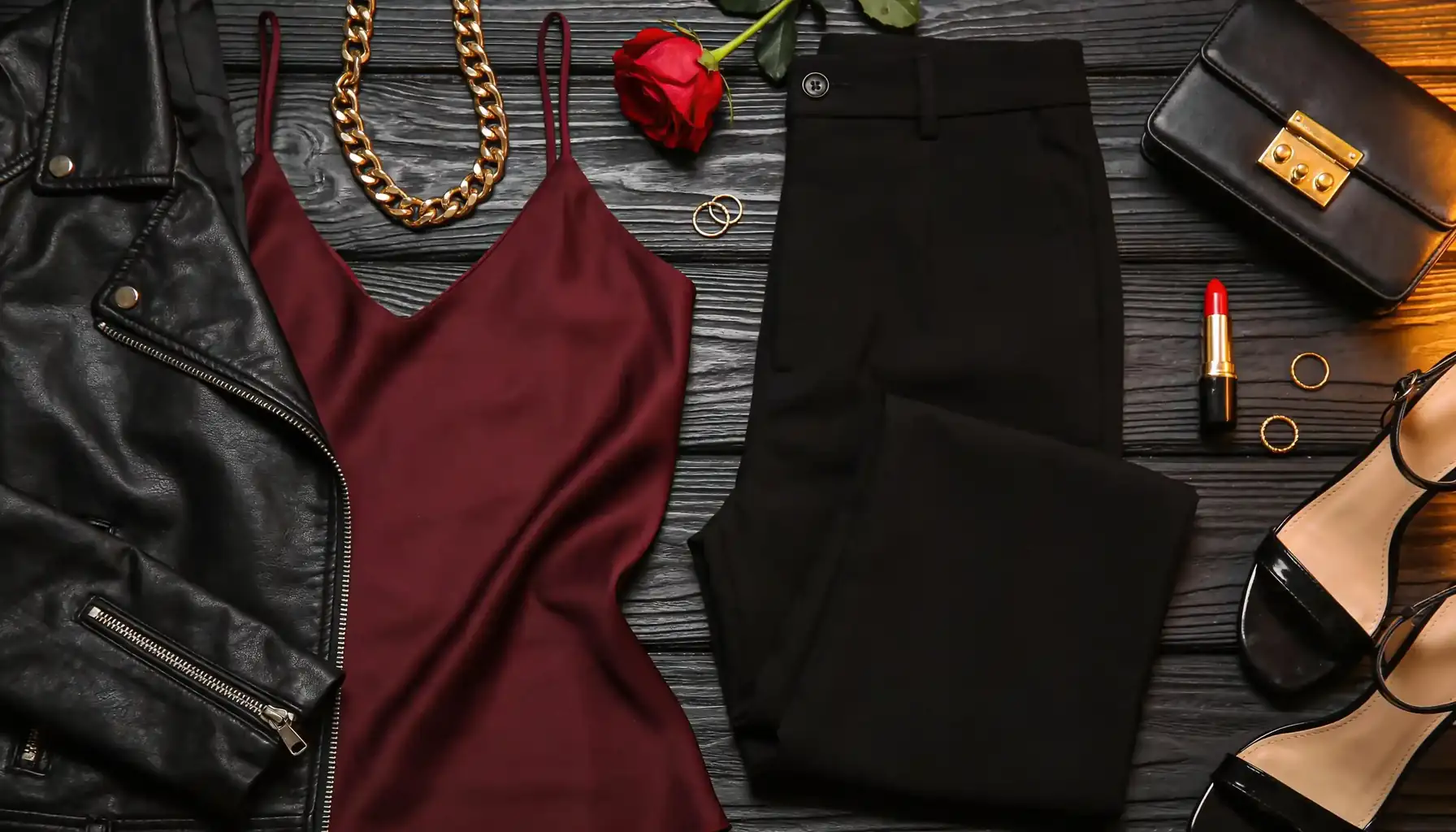Date night outfit flat lay with a black leather biker jacket, burgundy silk camisole, black tailored trousers, strappy heeled sandals, gold chain necklace, clutch bag, and red rose on dark surface