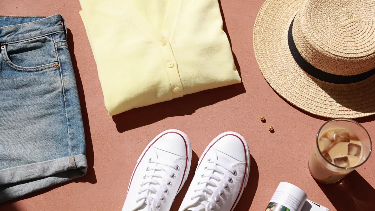 Butter yellow cardigan, washed denim shorts, white sneakers, gold studs, an iced coffee, and a magazine on pale terracotta for a relaxed weekend work session