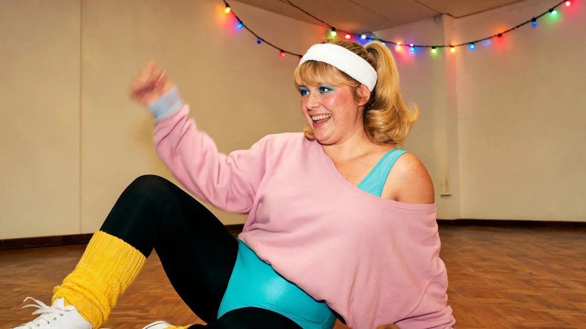 Femme d'une quarantaine d'années en justaucorps turquoise, legging noir brillant, sweat rose tombant sur une épaule, jambières jaunes et bandeau éponge blanc, dansant dans une salle des fêtes