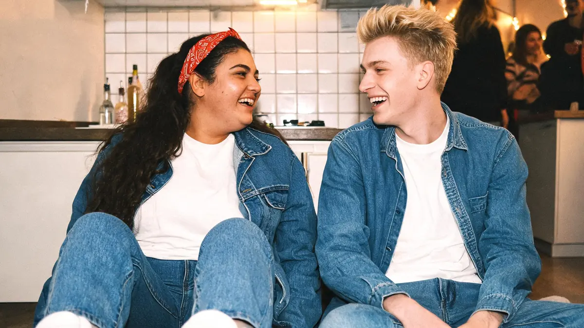 Couple d'une trentaine d'années entièrement en jean du même bleu, elle avec un bandana rouge dans les cheveux, lui avec une chemise en jean ouverte sur un t-shirt blanc, riant dans une cuisine pendant une soirée