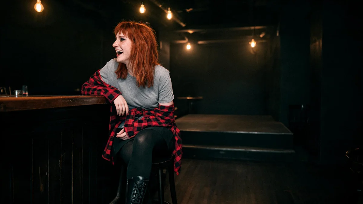 Femme rousse aux cheveux décoiffés en chemise à carreaux rouge nouée à la taille, t-shirt gris uni, jupe noire, collant opaque et rangers noires, accoudée au comptoir d'un petit bar