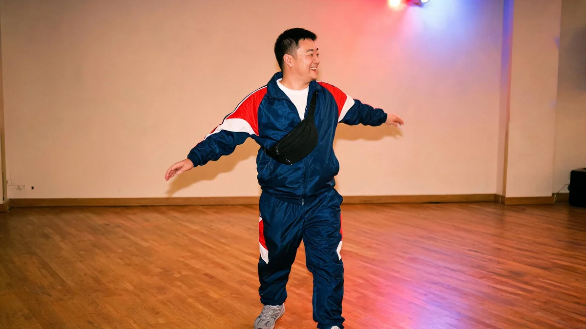 Homme d'une quarantaine d'années en survêtement complet en nylon bleu marine à empiècements rouges et blancs, t-shirt blanc et banane noire en bandoulière, dansant dans une salle des fêtes