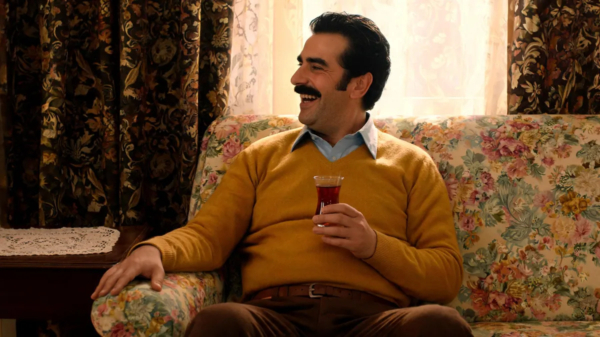 Gür siyah bıyıklı, yana taranmış saçlı orta yaşlı bir adam, açık mavi gömleğin üstüne hardal V yaka kazak ve kahverengi kumaş pantolonla çiçekli bir kanepede oturmuş, elinde ince belli çay bardağıyla gülüyor